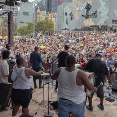 A live outdoor performance on a stage with a large crowd gathered at Federation Square.