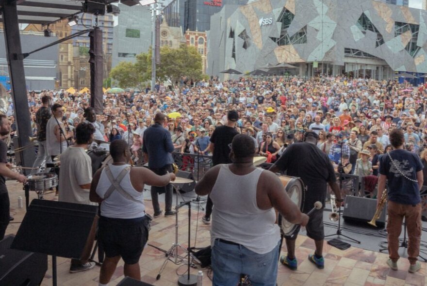 A live outdoor performance on a stage with a large crowd gathered at Federation Square.
