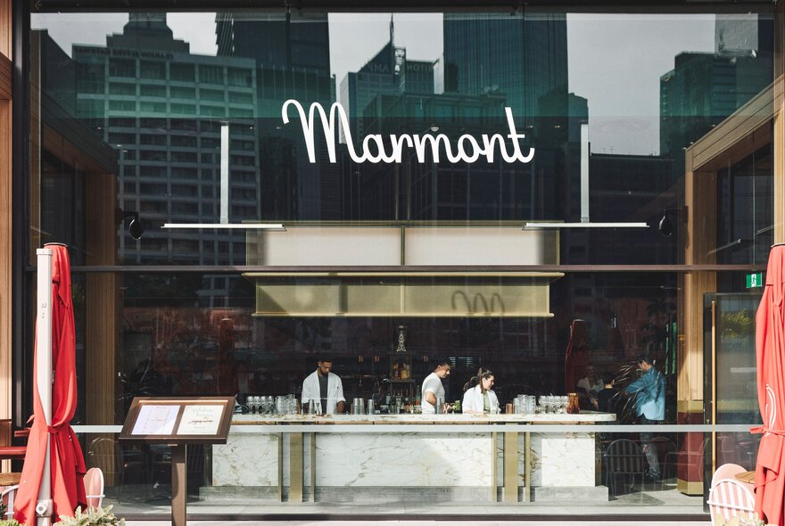 The view into a restaurant from the Yarra Riverwalk, showing a large central bar with staff, the words Marmont in cursive font displayed high on an external glass wall.