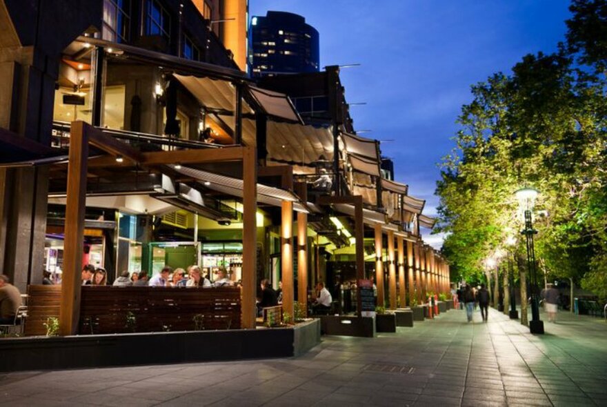 Exterior of BearBrass at twilight showing patrons seated in an outdoor area, and a colonnade of trees along a pedestrian walkway.