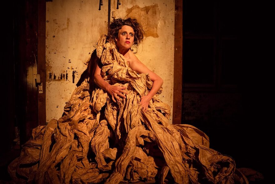 Performer dressed in rags, posed against a panel under a spotlight.