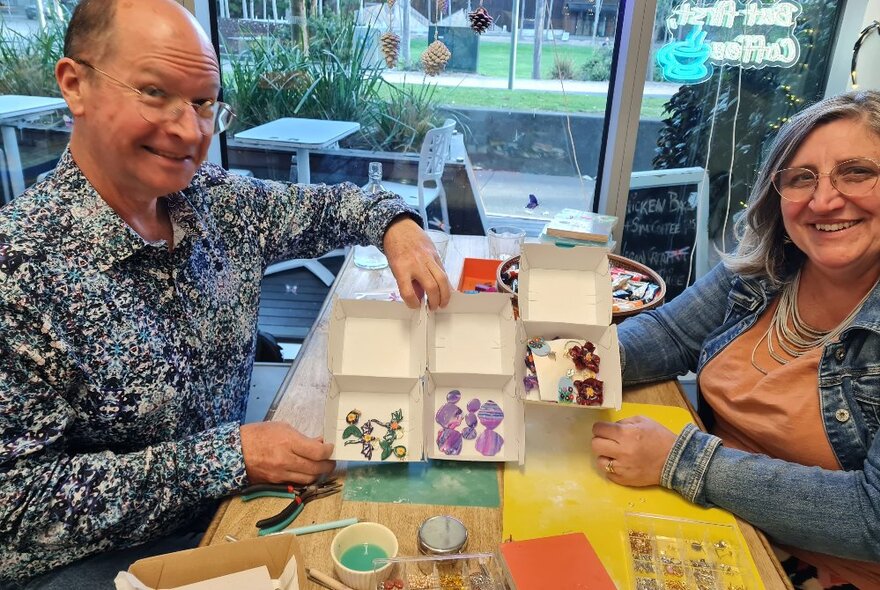 Two participants in a craft workshop seated at a table holding white trays with jewellery, with assorted crafts materials.