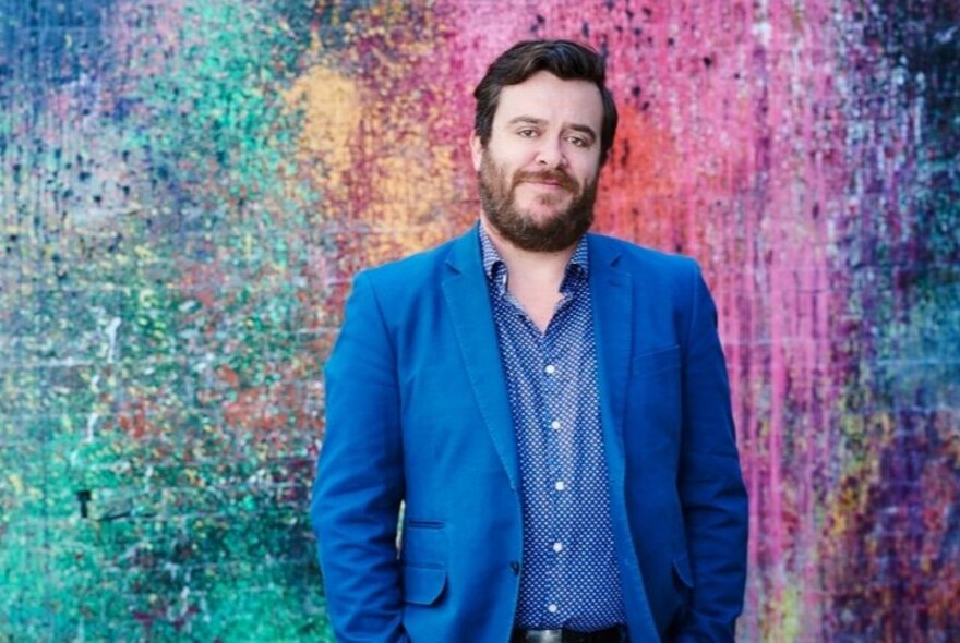 Michael Williams smiling with hands in pockets in front of a multicoloured wall. 