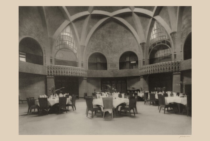 Historic photograph of a large, circular dining room of Newman College featuring a high, vaulted ceiling supported by intersecting architectural arches. Rounded tables draped in white tablecloths and surrounded by dark wooden chairs are arranged across the spacious floor.