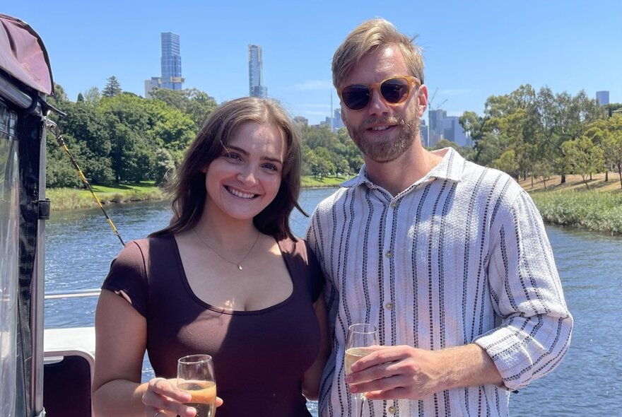 A smiling coupe standing up on a cruise boat, with Melbourne city buildings and Yarra River in the background.