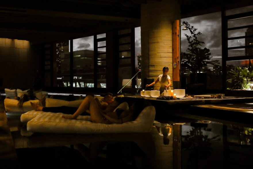 The dimly lit indoor swimming pool area at 1 Hotel Melbourne, with people relaxing on inflatables on the water and a person near the water's edge using bowls to create sounds. 