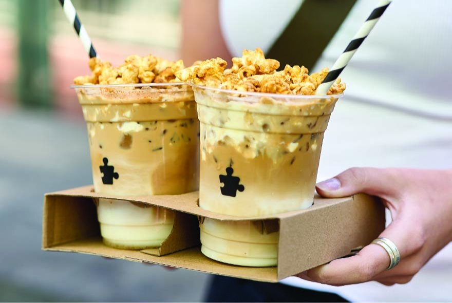 Two flavoured coffee drinks in clear plastic cups with straws in a cardboard serving tray held by hands. 