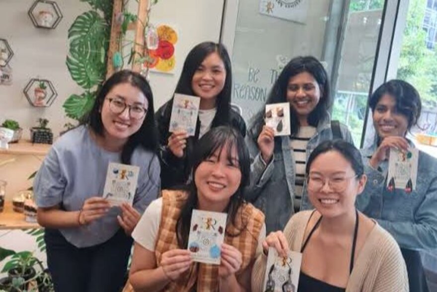 Six smiling workshop participants in a studio space, proudly holding up jewellery they have made.