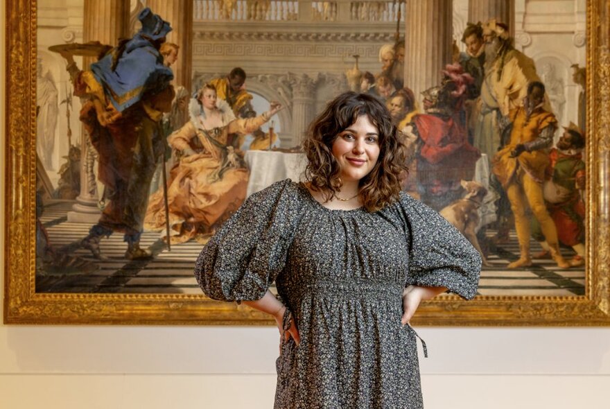 Author and cook Julia Busuttil Nishimura standing with her hands on her hips, smiling, in front of a large framed Baroque oil painting at the National Gallery of Victoria.