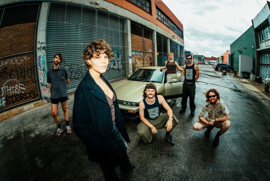 A six-piece band posing for a photo in a laneway with a car, and industrial buildings.