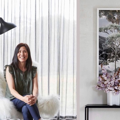 A smiling woman sitting in a fluffy white armchair with her legs crossed and her hands resting on her knees; there is a white curtain. an artwork and a vase of flowers behind her.