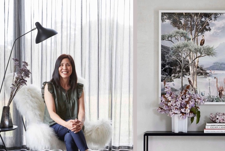 A smiling woman sitting in a fluffy white armchair with her legs crossed and her hands resting on her knees; there is a white curtain. an artwork and a vase of flowers behind her.