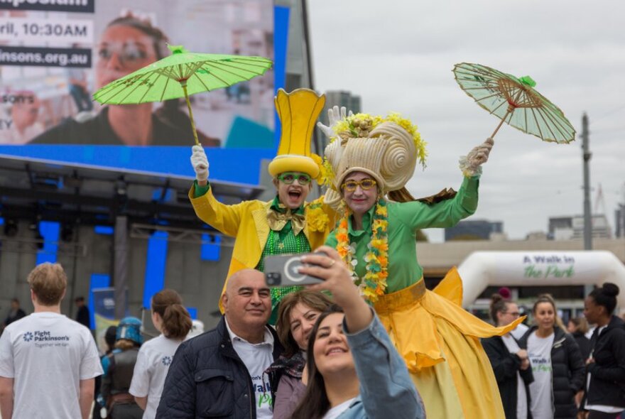 Two performers on stilts wearing yellow and green costumes with parasols, with a young woman taking a selfie in front of them with her parents.