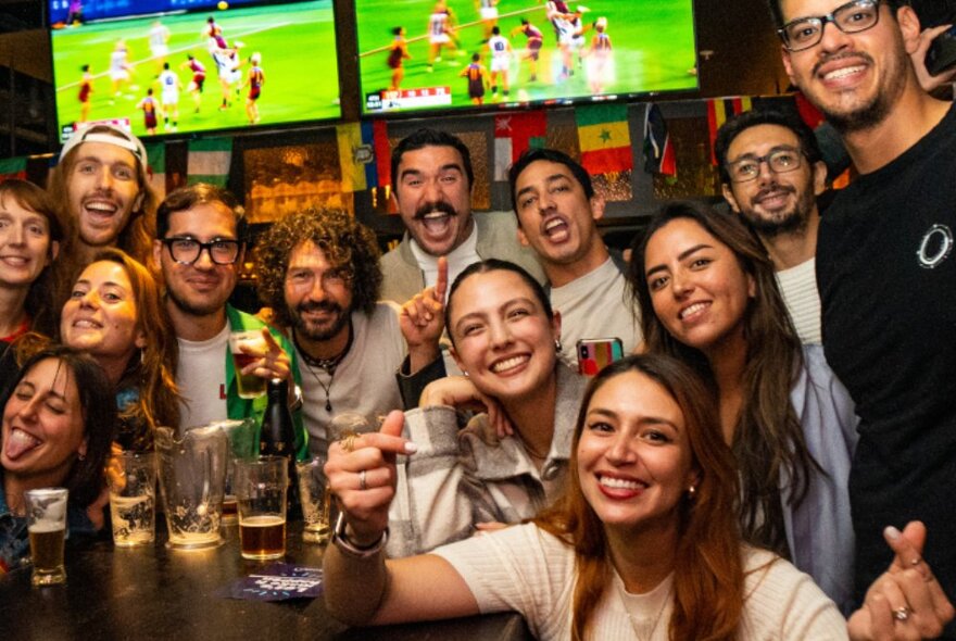 A very happy group of people in a bar posing for a photo with sport on the TVs in the background.