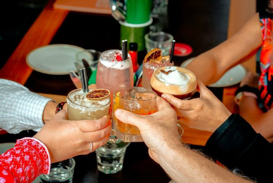 Five hands holding different cocktails in glasses, bringing them all together above a table in a celebratory clinking glasses cheers gesture.