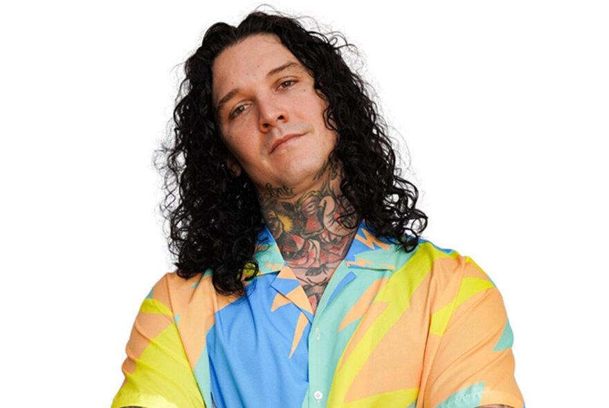 Artist Josh Deane, with long dark hair, wearing an open necked brightly coloured shirt, his neck adorned with tattoo designs.