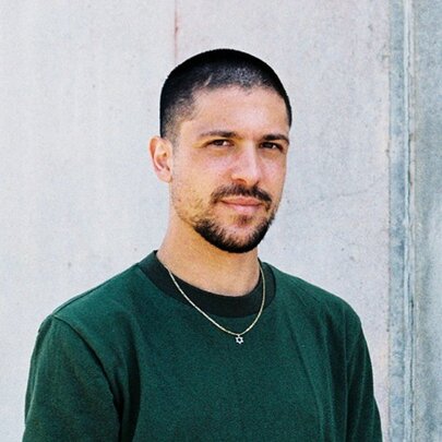 A portrait of Joshua Moshe,a young man with short dark hair and a closely cropped beard, looking at the camera, wearing a dark green top.