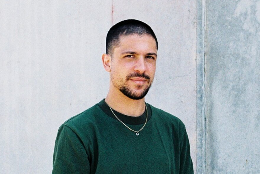 A portrait of Joshua Moshe,a young man with short dark hair and a closely cropped beard, looking at the camera, wearing a dark green top.