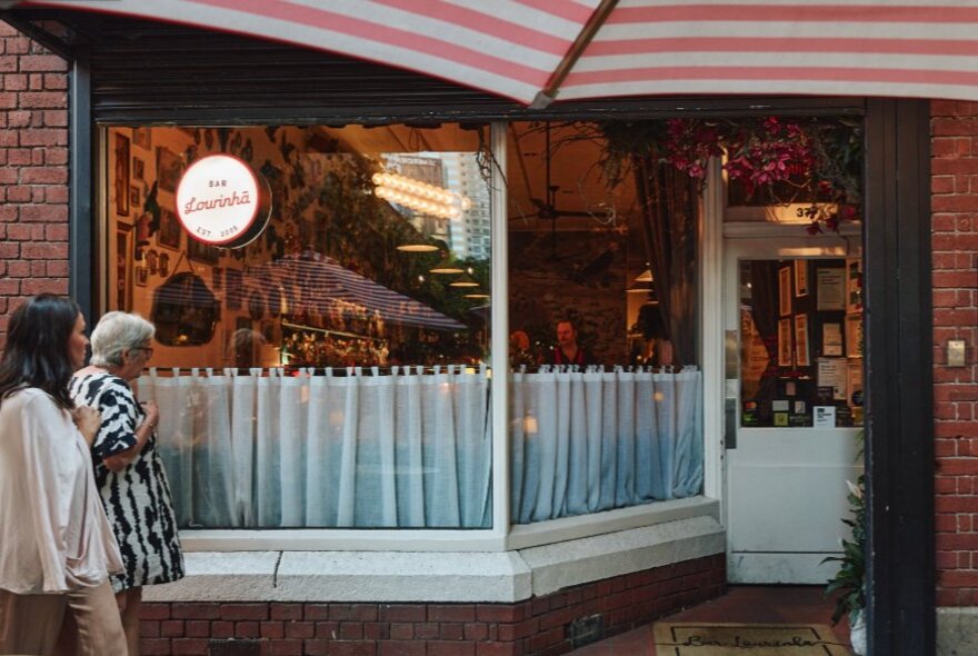 Two people walking past a bar with window curtains, striped umbrella and windows.