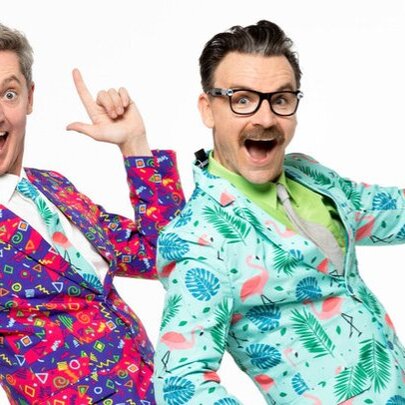 Two men in brightly patterned suits leaning back with one index finger in the air and mouths open in surprise. 