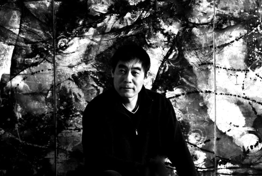 Artist Zhou Xiaoping, seated in front of one his his large artworks; black and white image.