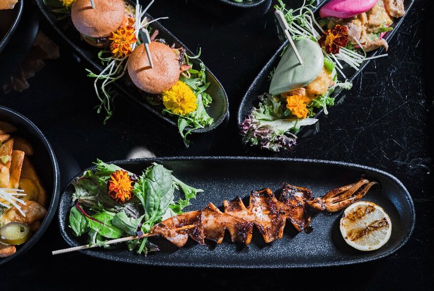 Looking down on black long thin plates of small bites or bao, sliders and squid on a stick with grilled lime.