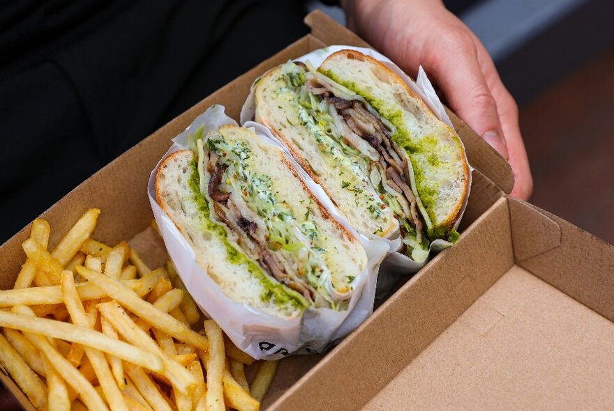 Generously-filled sandwich next to chips in a cardboard container, hand visible. 