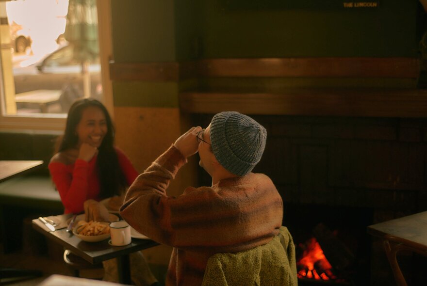Two friends seated across from each other at an indoor table at The Lincoln, laughing and enjoying their meal in front of an open fire.