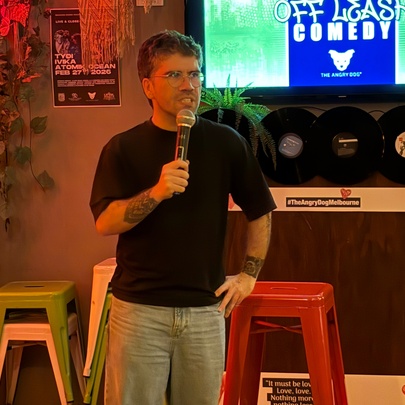 A man in a black t-shirt standing with a microphone in front of a presentation screen and two stools, inside a bar setting. 