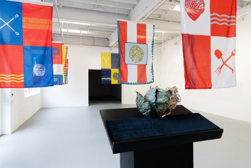 Flags hanging from the ceiling in a gallery room, images of cabbages and gardening tools printed on them, and a sculpture of a cabbage on a black plinth display in the foreground.