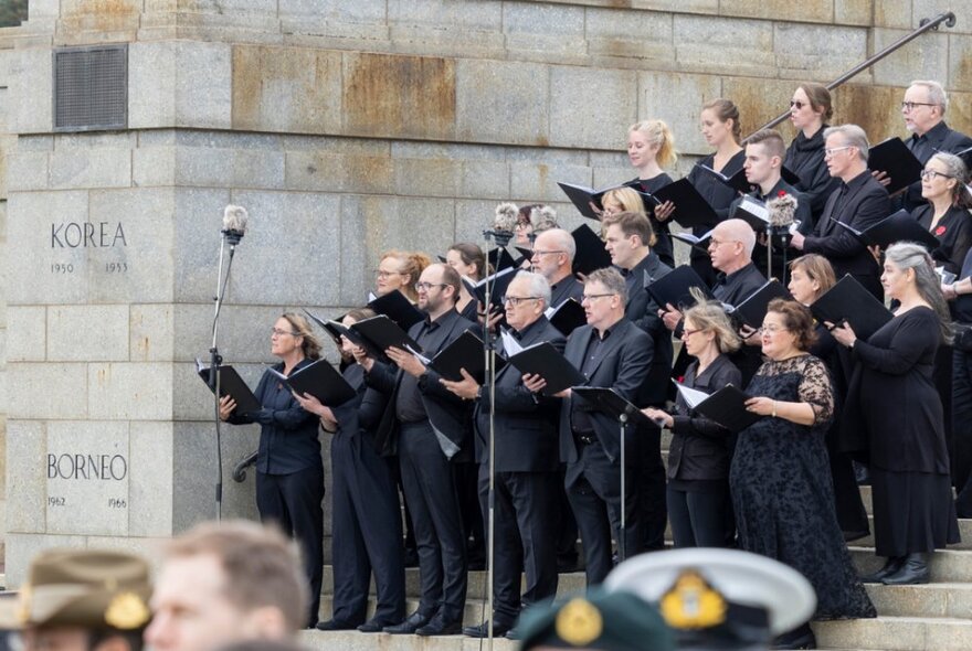 A choir dressed in black, standing on the steps of the Shrine of Remembrance in Melbourne, each with songbooks in their hand.