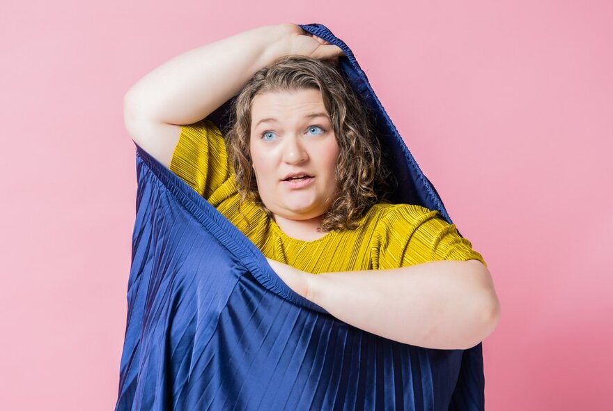 A woman putting herself into a blue silky bag against a pink background. 