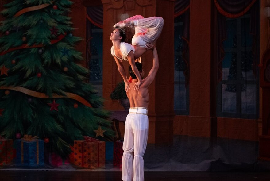 A shirtless male dancer holding a dancer above his head who balances her arms on his shoulders while bending her body into a curve so that her toes are touching the top of her head.