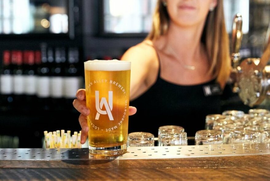 Blonde waitress in a black singlet standing behind the bar placing a freshly poured beer on the bar top.