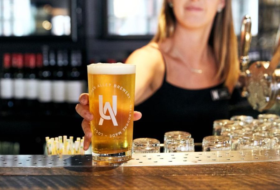 Blonde waitress in a black singlet standing behind the bar placing a freshly poured beer on the bar top.