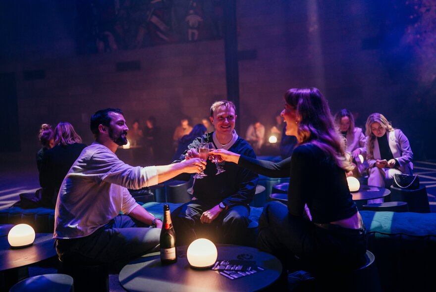 Three people clinking glasses in a darkened room with small glowing lights. 