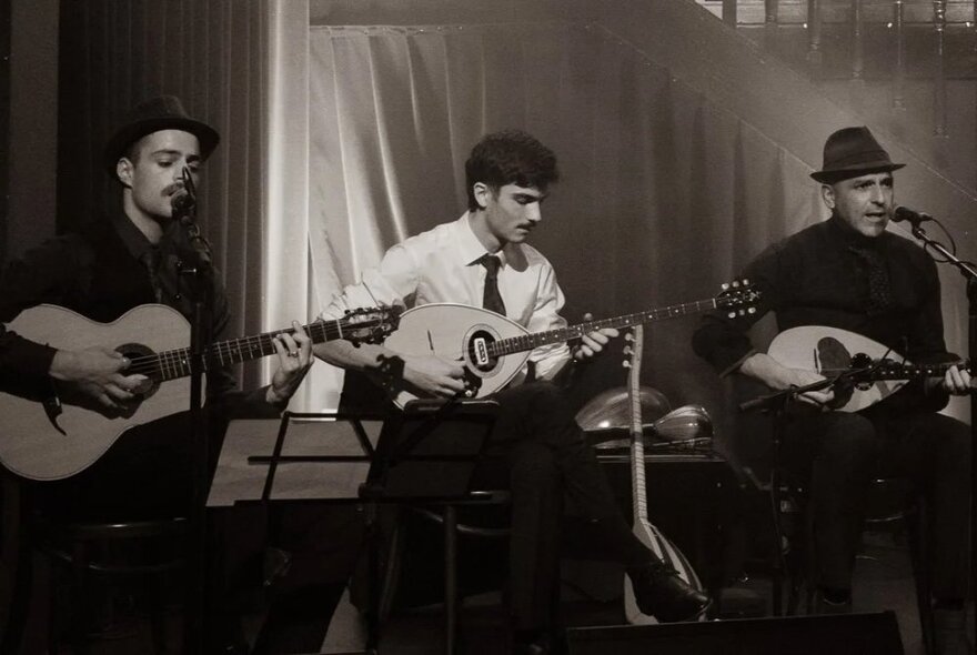 Three musicians seated in front of music stands and microphones playing guitar and bouzoukis.