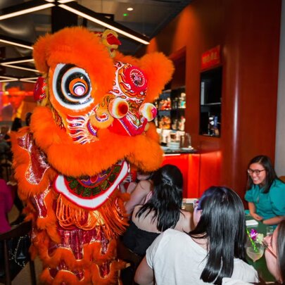 A lion dancer in a large red suit performing in a busy restaurant.