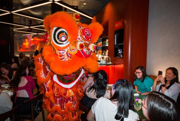 A lion dancer in a large red suit performing in a busy restaurant.