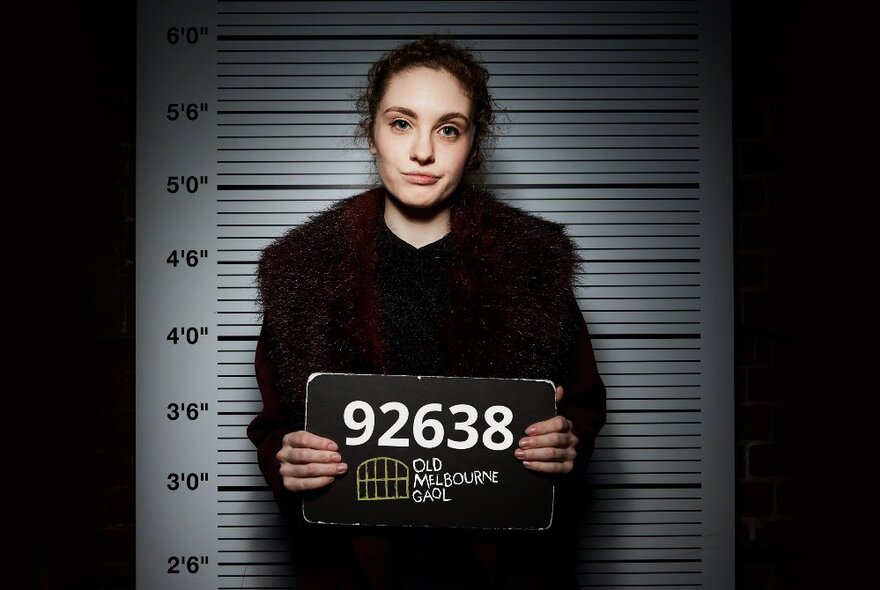 Woman holding a criminal identity number, standing in front of a wall height measurement screen, looking nonplussed.