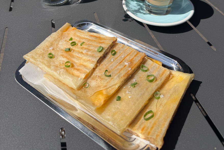 A high-angle shot of several pieces of golden-brown, folded flatbread garnished with sliced spring onion greens, served on a rectangular metal tray on a dark outdoor table.