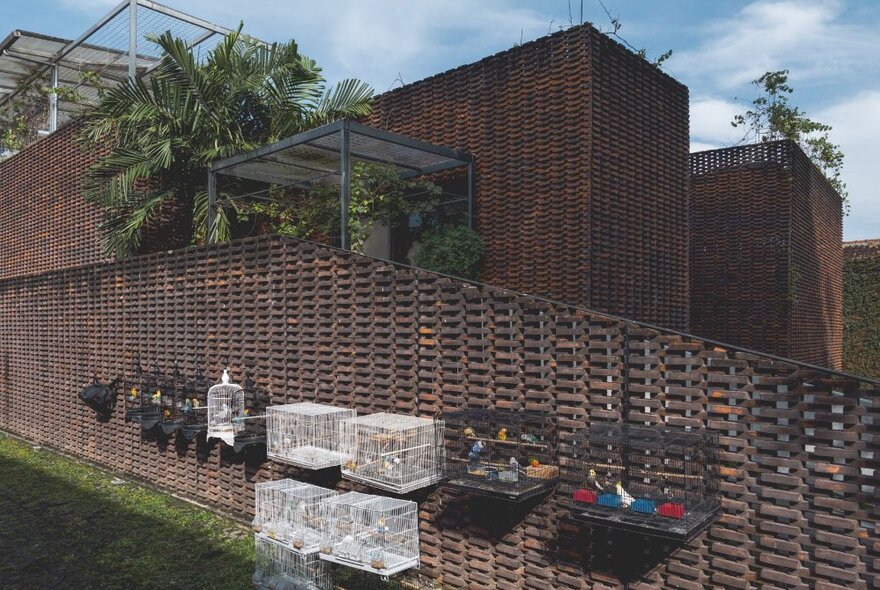 Building exterior with walls that appear woven, bird cages hanging off the external fence, with some greenery between the fence and building.