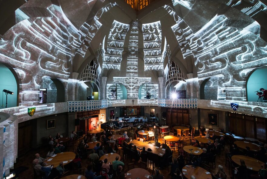 Interior shot of a large, historic dining hall with a soaring vaulted ceiling illuminated by white patterned light projections, with people seated at tables below.