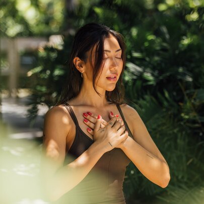Yoga practitioner with crossed hands on her upper chest, her eyes closed, in a leafy setting.
