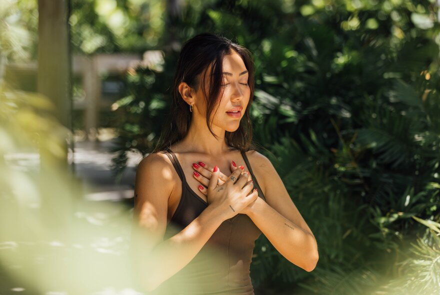 Yoga practitioner with crossed hands on her upper chest, her eyes closed, in a leafy setting.