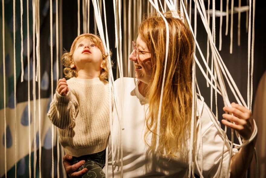 A mother and her child experiencing an artplay installation where many cords are suspended from the ceiling, mimicking rain.