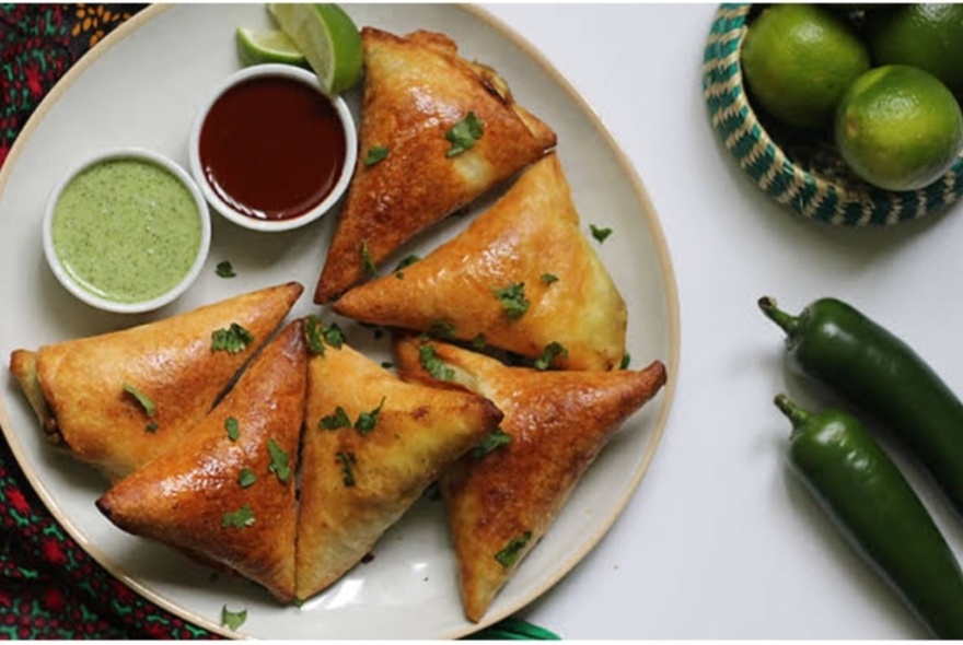 Triangular samosas on a plate with two dipping sauces and chillies and bowl of limes nearby