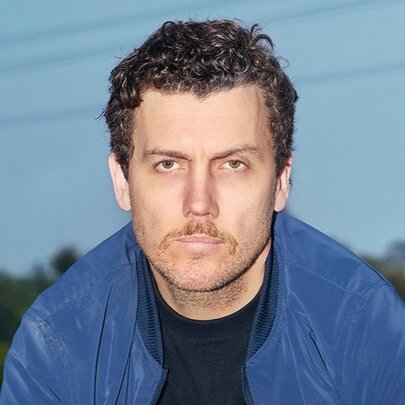 Stand-up comedian Daniel Connell, a man in his 30s with short brown hair and stubble on his chin, with a neutral expression, wearing a blue jacket over a black t-shirt.