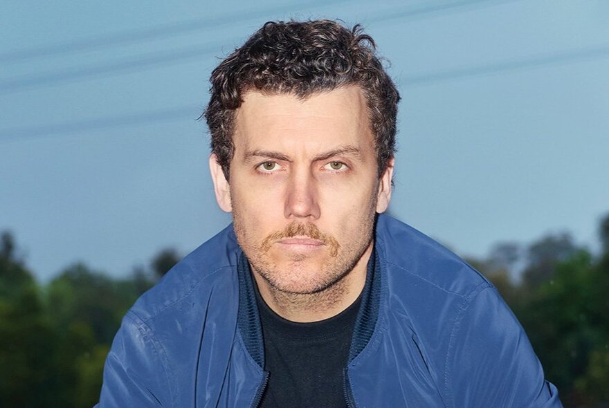 Stand-up comedian Daniel Connell, a man in his 30s with short brown hair and stubble on his chin, with a neutral expression, wearing a blue jacket over a black t-shirt.