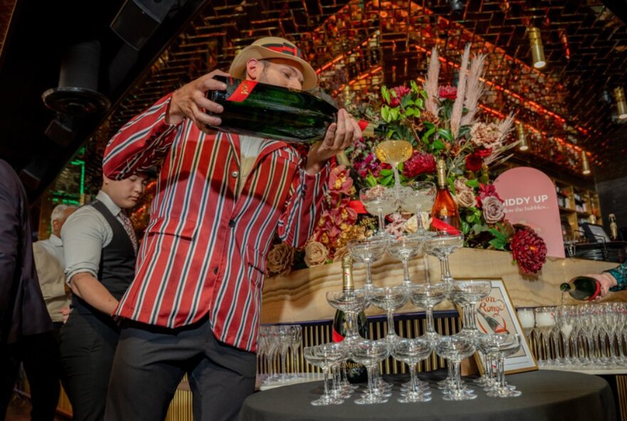 A waiter in a red-striped jacket pouring a magnum of champagne into a champagne tower in front of a floral arrangement of roses.